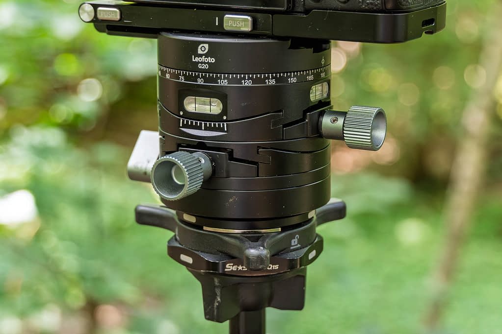 Close-up of a black Leofoto G20 geared tripod head, an excellent compact choice among the best tripod heads for woodland photography. Two large adjustment knobs, measurement markings, and locking mechanisms stand out against blurred outdoor greenery.