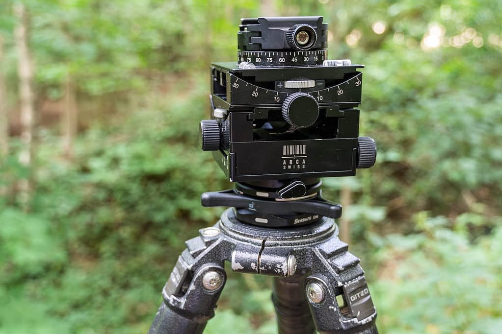 A close-up of an Arca Swiss C1 Cube geared tripod head with adjustment knobs and measurement markings, mounted on a sturdy Gitzo tripod - one of the best tripod setups for woodland photography. The background consists of blurred green foliage and trees, indicating an outdoor forest setting.