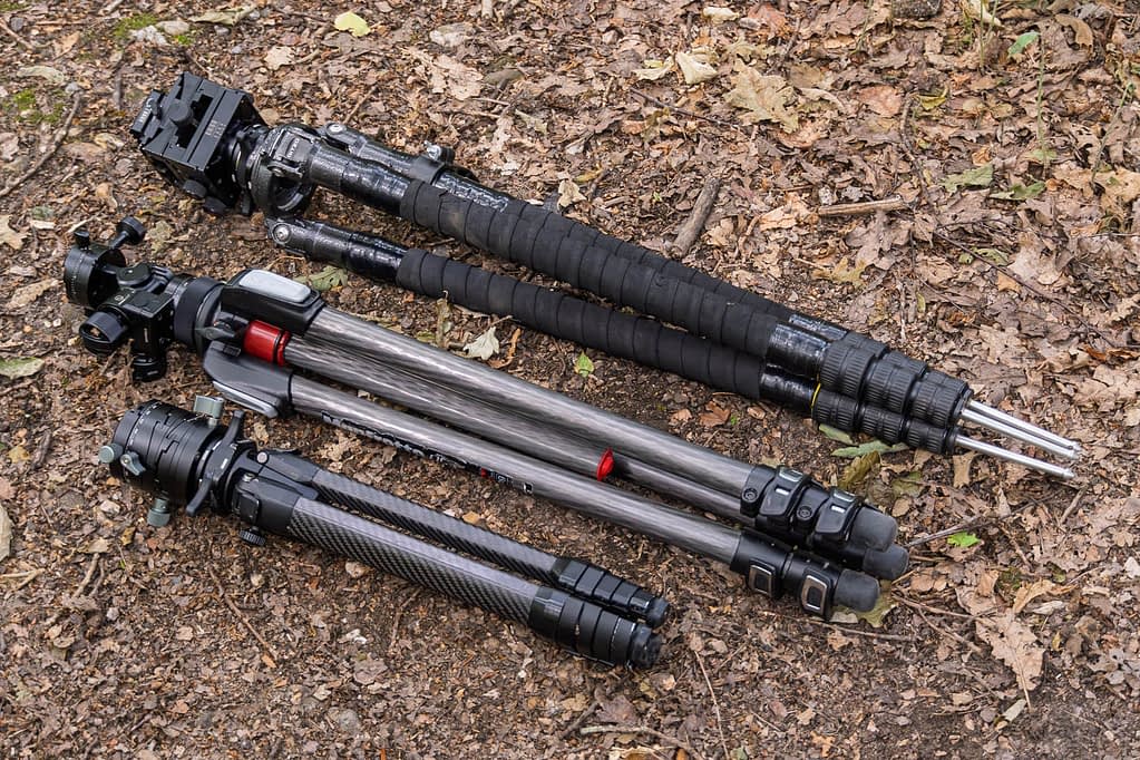 Three different camera tripods - the Gitzo GT3541XLS, the Manfrotto 055CXPRO3 and the Peak Design Travel Tripod - regarded as some of the best tripods for woodland photography, lie parallel on a forest floor strewn with dry leaves and twigs. Each features a distinctive design and colour, with visible legs, locking mechanisms, and head attachments.