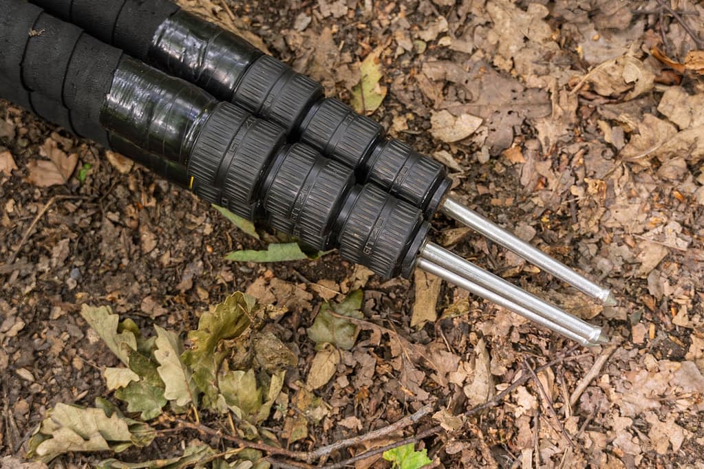 Two black telescopic tripod legs with rubber grips and twist locks are extended over dry soil and scattered brown leaves—an ideal setup for anyone seeking the best tripod for woodland photography among twigs, debris, and leaf litter.