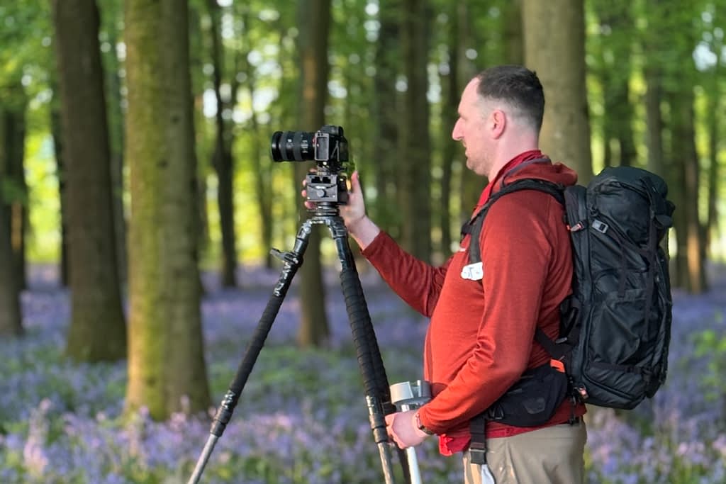 Professional Woodland Photographer Tim Smalley adjusts his camera on a tripod in a forest filled with tall trees and blooming bluebells, as soft sunlight filters through the greenery.
