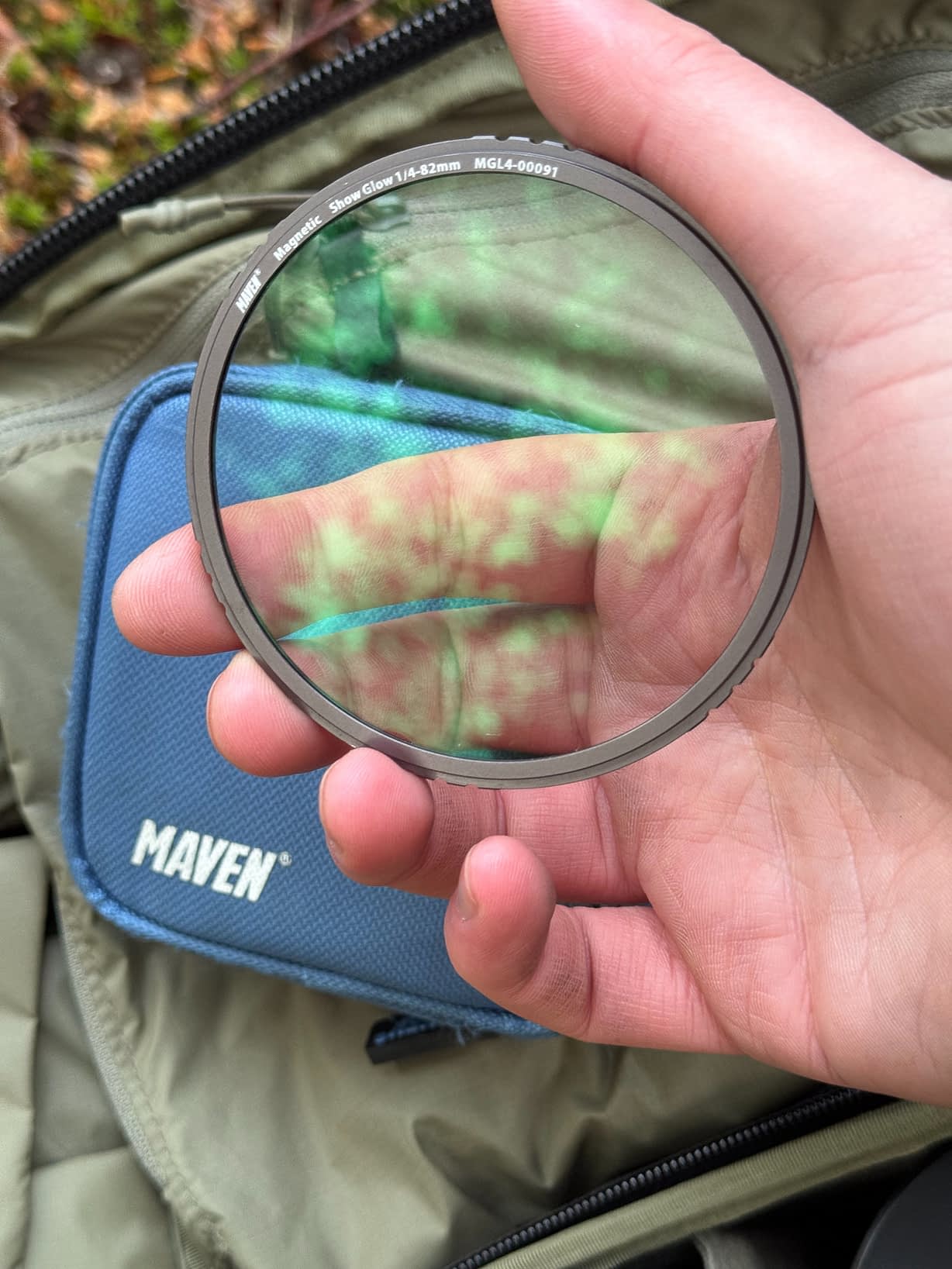 A hand holds a magnetic black mist filter with a slim metal rim, reflecting green tones. Behind, a blue MAVEN pouch sits on olive-green fabric.