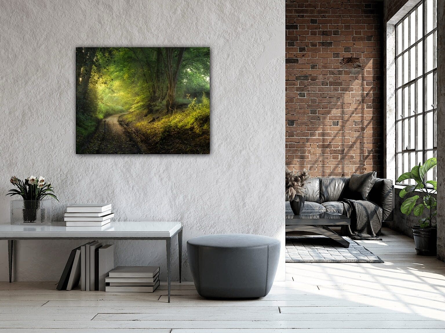 IMG_2182 A contemporary living room featuring large windows, exposed brickwork, and indoor plants. By the windows is a black sofa with cushions. A light-coloured desk holds books and flowers, while a painting of a forest path hangs on a white wall above a grey ottoman.