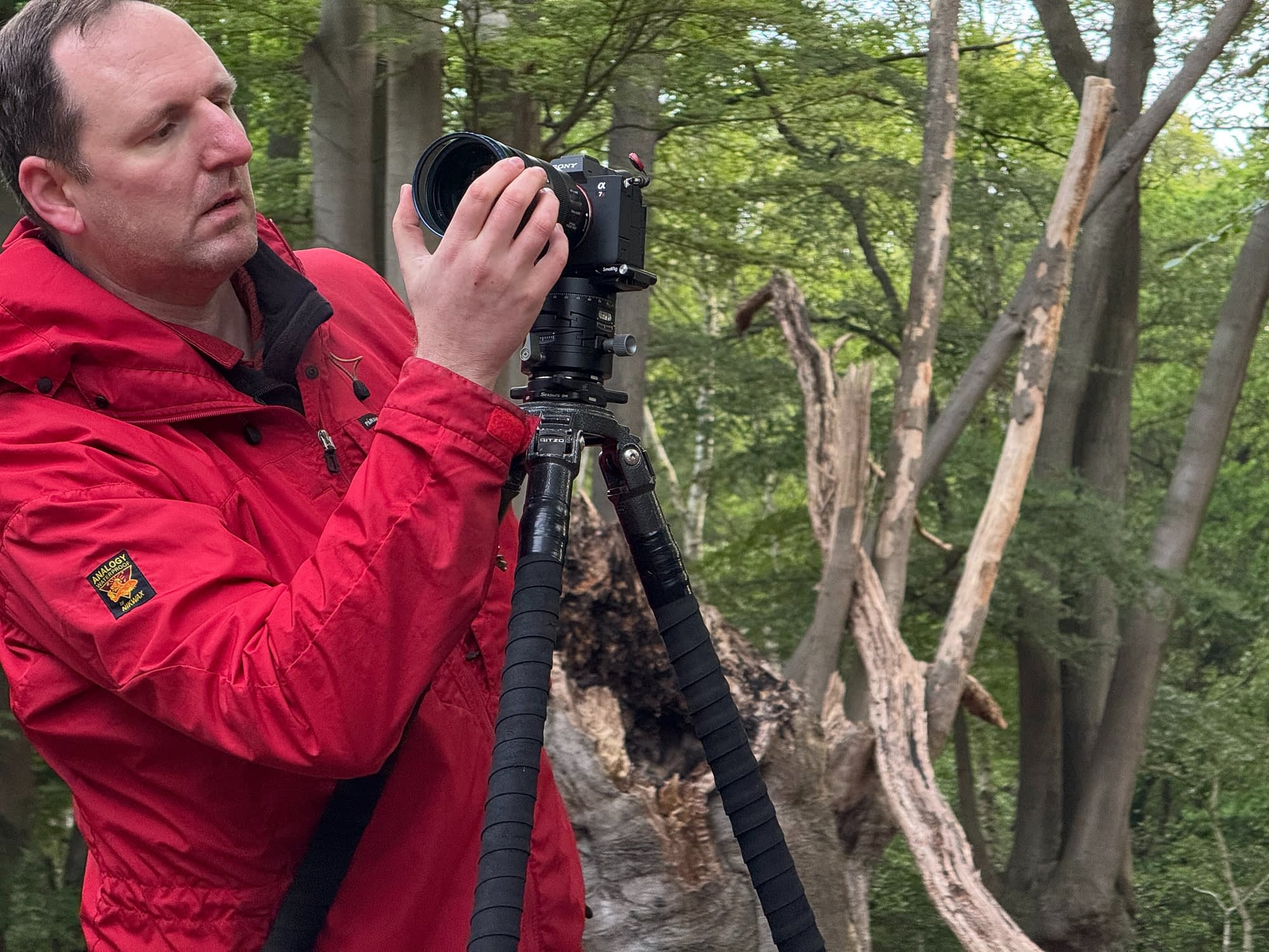 Tim Smalley adjusts a camera on a tripod in a verdant forest, focused on settings amid green trees for nature photography.