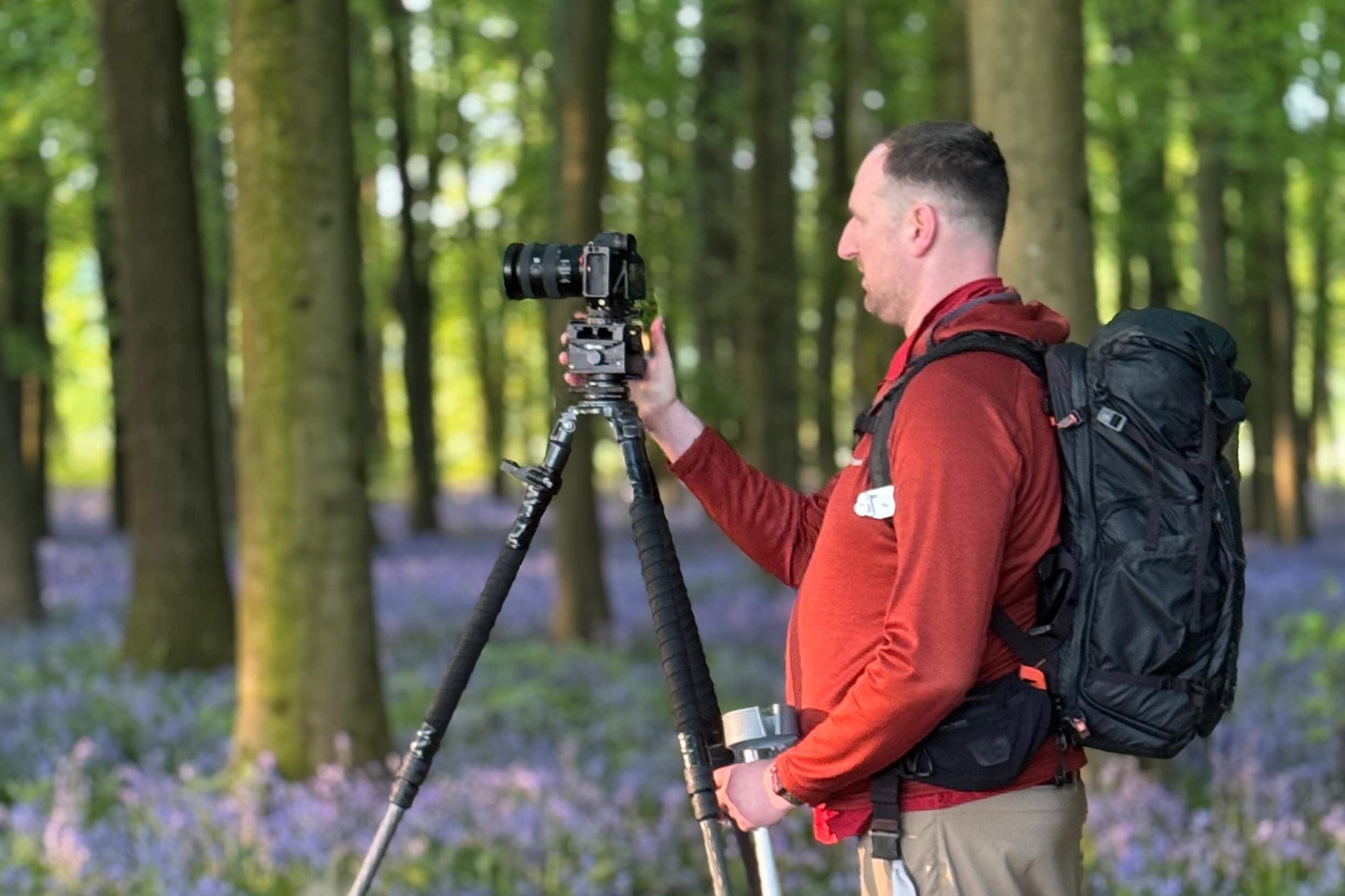 IMG_7341 Professional Woodland Photographer Tim Smalley adjusts his camera on a tripod in a forest filled with tall trees and blooming bluebells, as soft sunlight filters through the greenery.